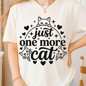 May include: A cream-coloured short-sleeve t-shirt featuring a black graphic print. The design includes three cat illustrations, several small heart shapes, and star accents, with the text "just one more cat" centred in the middle.