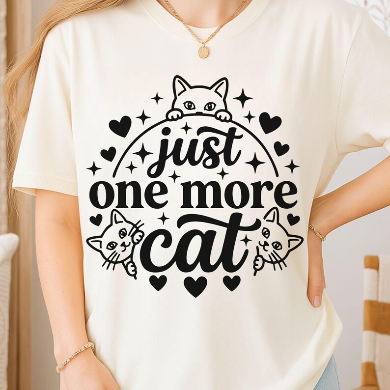 May include: A cream-coloured short-sleeve t-shirt featuring a black graphic print. The design includes three cat illustrations, several small heart shapes, and star accents, with the text "just one more cat" centred in the middle.