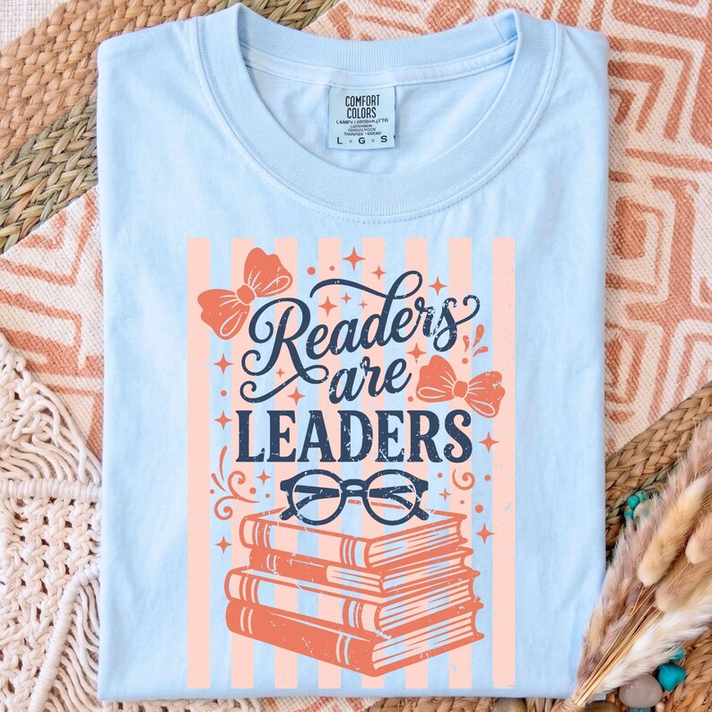 Op de afbeelding: Lichtblauw T-shirt met de tekst "Readers are Leaders" in een decoratief lettertype, met strikken, een bril en een stapel boeken. Het ontwerp is in koraal en marineblauw, met verticale strepen.