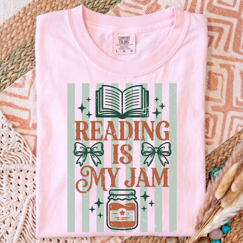Op de afbeelding: Lichtroze t-shirt met een grafisch ontwerp. Het ontwerp bevat een open boek, strikken, een pot jam en de tekst "READING IS MY JAM" in groen en bruin. Het shirt heeft een ronde hals en korte mouwen.