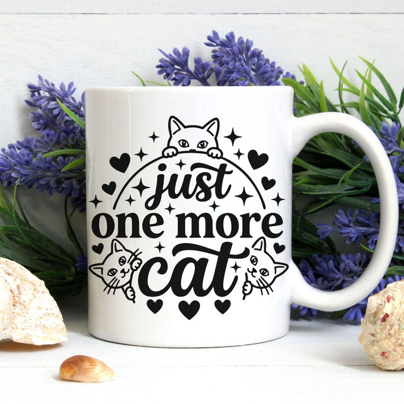 May include: A white ceramic mug featuring a black graphic design of three cat faces, small hearts, and stars. The text "just one more cat" is printed in the centre in a bold, stylised font. The mug has a curved handle on the right side.
