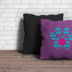 May include: A square purple throw pillow featuring a bright blue chevron-patterned paw print graphic with a circular hot pink monogram center containing the letters AAB, positioned in front of a solid black square throw pillow.