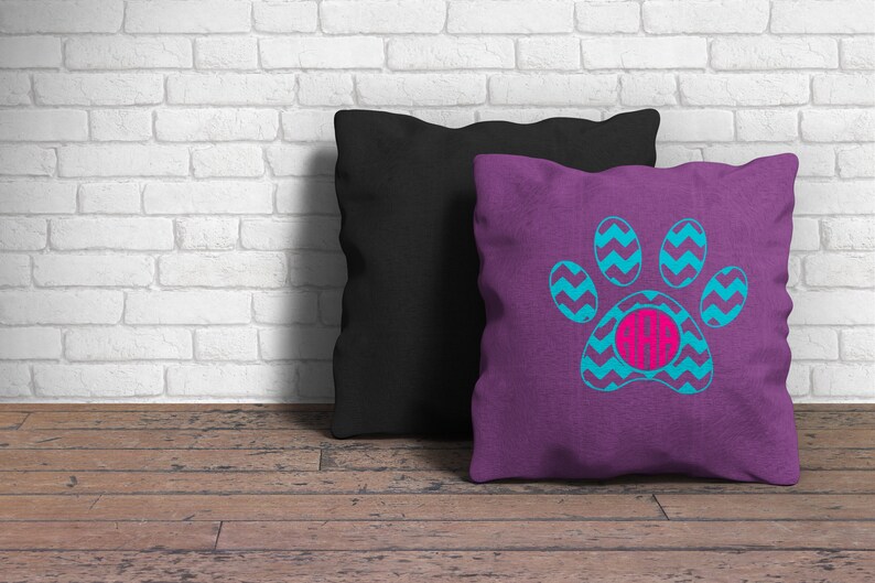 May include: A square purple throw pillow featuring a bright blue chevron-patterned paw print graphic with a circular hot pink monogram center containing the letters AAB, positioned in front of a solid black square throw pillow.