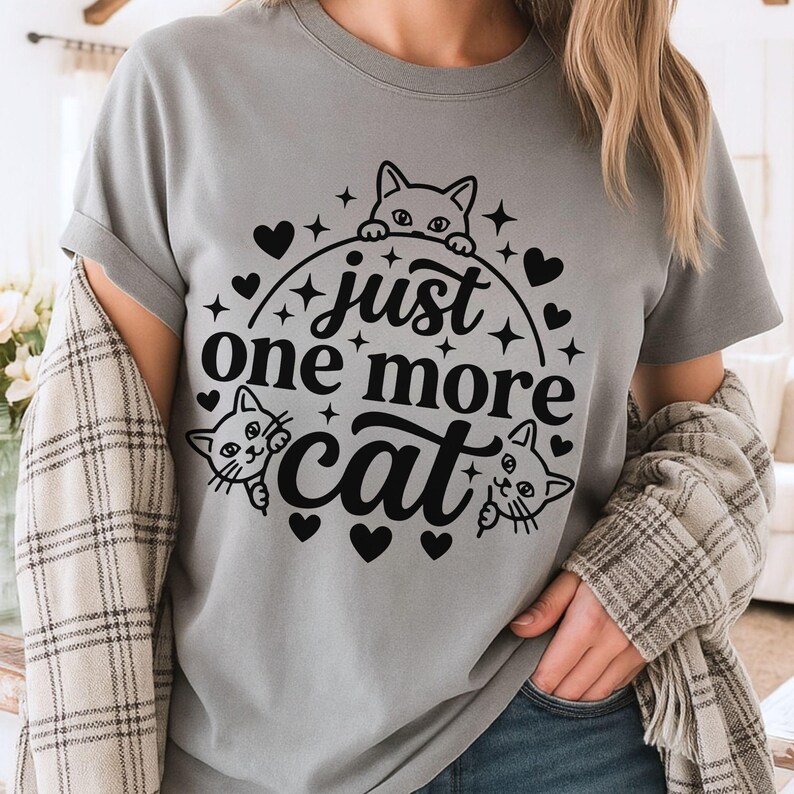 May include: A grey t-shirt featuring a black graphic print of three cat faces peeking out from behind a circular border. The text "just one more cat" is centred within the circle, surrounded by small black hearts and star-shaped accents.