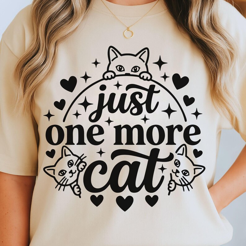 May include: A beige crewneck t-shirt featuring a black graphic print of three cats and the text "just one more cat" surrounded by small heart and star shapes. The design is centred on the chest of the garment, which has short sleeves.