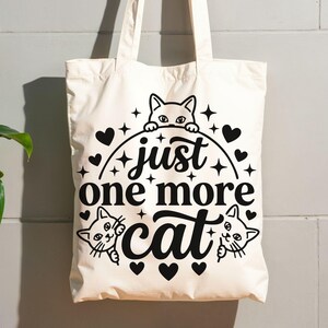 May include: A cream-coloured canvas tote bag featuring a black graphic print. The design includes three cat faces peeking out, surrounded by small hearts and star shapes, with the text "just one more cat" centred in a bold, decorative script font.