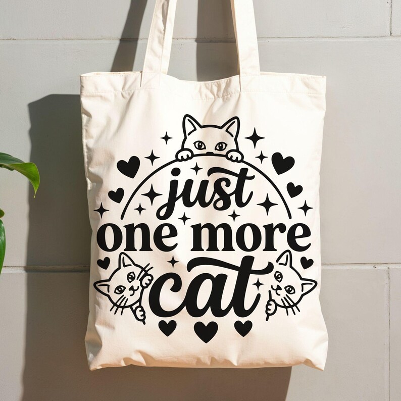 May include: A cream-coloured canvas tote bag featuring a black graphic print. The design includes three cat faces peeking out, surrounded by small hearts and star shapes, with the text "just one more cat" centred in a bold, decorative script font.