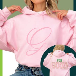 Puede incluir: Una sudadera con capucha rosa claro con una gran "G" en cursiva en la parte delantera. La parte trasera de la sudadera con capucha presenta las palabras "FOR GOOD" en una fuente decorativa. El modelo lleva vaqueros azules.