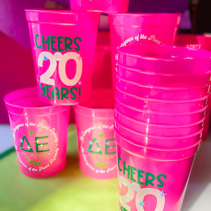 May include: Bright pink plastic cups with text that reads "CHEERS to 20 YEARS!" and "ΔE". Some cups have additional text: "The Emergence of the Pearl Dynasty" and "EST SPR 2004+". The cups are stacked and arranged on a colorful background.