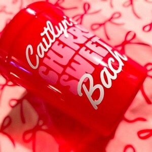 May include: A red tumbler with the text "Caitlyn's Sweet Bach" in white and pink lettering. The tumbler is on a pink surface with a pattern of red bows.
