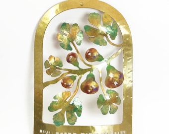 May include: Gold metal wall hanging with a cut-out design of a fig tree with green leaves and purple figs. The Hebrew text "ברוך אתה ה' אלוהינו מלך העולם" is inscribed at the bottom.