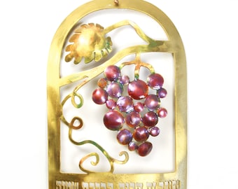 May include: A gold-colored metal mezuzah with a grapevine design and a cluster of purple grapes. The Hebrew text "Zeh Kevod Ba'aseh She'avti" is inscribed on the bottom.