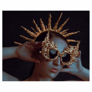 May include: A woman wearing a gold crown with a pointed design and a pair of ornate gold sunglasses with a floral pattern.