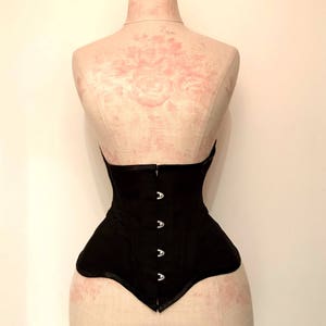 May include: A black corset with silver metal clasps on a white mannequin. The corset has a smooth, satin-like finish and a pointed bottom.