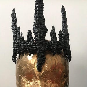 May include: A black crown with a jagged, dripping design. The crown is made of a textured material and has a sharp, pointed shape.