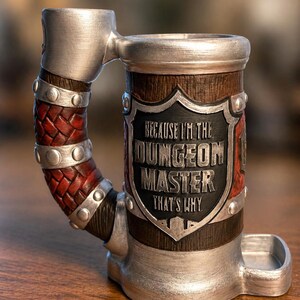 May include: A cylindrical mug with a metallic silver finish, featuring a handle wrapped in red braided material with silver studs. A central black shield plaque displays the raised silver text: BECAUSE I'M THE DUNGEON MASTER THAT'S WHY.