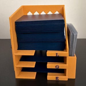 May include: An orange plastic card organizer featuring three horizontal shelves labeled with the letters L, G, and E in purple. The organizer holds a large stack of dark blue cards on the top shelf and a smaller stack of clear-sleeved cards on the side.