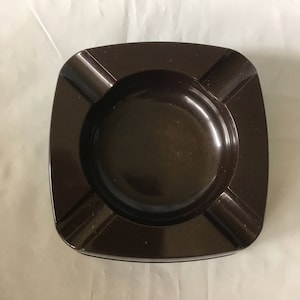 May include: A dark brown square ashtray with four indentations for cigarettes.