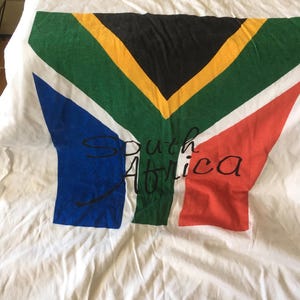 Pode incluir: Camiseta branca com um gr&aacute;fico da bandeira da &Aacute;frica do Sul e o texto "South Africa" impresso em preto.