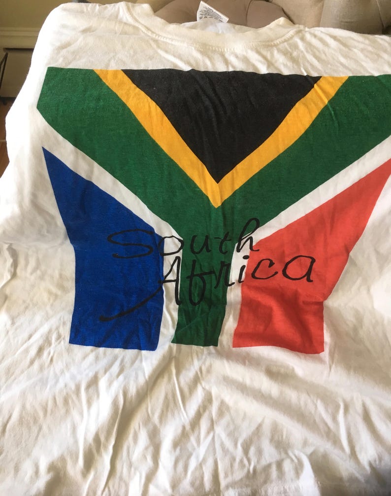 Pode incluir: Camiseta branca com um gr&aacute;fico da bandeira da &Aacute;frica do Sul e o texto "South Africa" impresso em preto.
