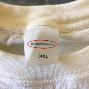 Pode incluir: Camiseta branca com uma etiqueta branca que diz "Escape" em letras azuis e "XXL" em letras pretas.
