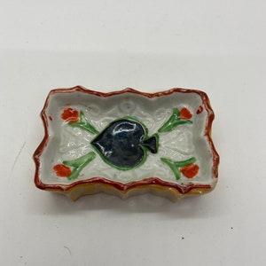 May include: A small, rectangular ceramic dish with a yellow rim and a red and green floral design. The center of the dish features a black spade playing card.