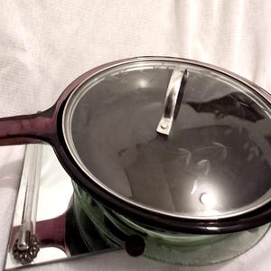 May include: A vintage glass saucepan with a clear lid and a long, purple handle. The pan sits on a mirrored stand with decorative accents. The glass is a light green color.