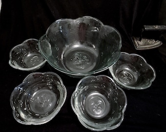 May include: A clear glass serving set consisting of one large bowl and four smaller matching bowls. Each piece features a scalloped edge and a molded floral pattern at the center of the base, with a smooth, transparent finish throughout.