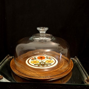 May include: A vintage cheese dome with a clear glass cover and a wooden base. The base features a decorative floral design in orange, yellow, and green. The dome sits on a mirrored tray, creating a classic presentation.