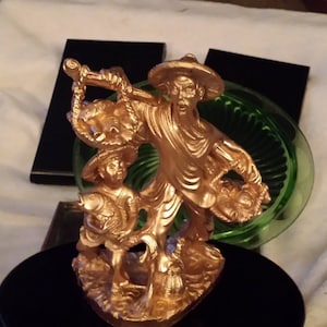 May include: A gold-colored figurine of a fisherman holding a fish and a basket of fish. The figurine is standing on a base with a wave design. The figurine is on a black surface.
