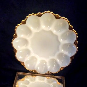 May include: A white, scalloped-edge serving dish with a gold-colored trim. The dish has a central circular area surrounded by 12 rounded indentations. The dish is likely made of glass or ceramic and is displayed on a dark blue surface.