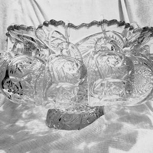 May include: A clear, cut-glass bowl with a scalloped rim and intricate geometric patterns. The bowl is supported by a pedestal base and features a series of attached, rounded decorative elements. The bowl is likely used for serving or display.