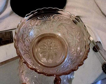 Può includere: Una ciotola di vetro trasparente, tinta di rosa, con bordo smerlato. La ciotola presenta un motivo floreale in rilievo, con fiori e foglie. La ciotola è appoggiata su una superficie riflettente.