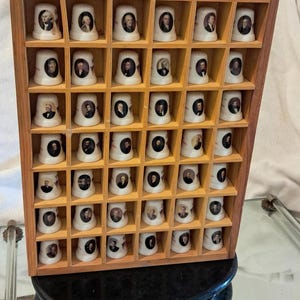 May include: A wooden display case filled with 48 white porcelain thimbles, each featuring a portrait. The thimbles are arranged in a grid pattern within the case, which sits on a black, oval-shaped base.