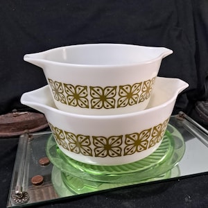 May include: Two white mixing bowls with a green floral pattern. The bowls are nested together and are sitting on a green glass surface.