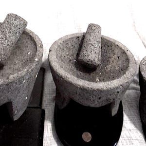 May include: Three grey volcanic stone molcajetes of varying sizes, each featuring a rough, porous texture, a circular basin, and three short legs. Each mortar is accompanied by a matching cylindrical stone pestle, known as a tejolote, resting inside.