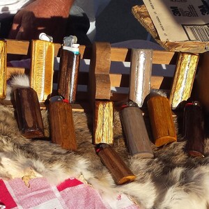 Puede incluir: Una colecci&oacute;n de estuches para encendedores de madera con varios patrones de veta de madera y acabados. Algunos estuches tienen una incrustaci&oacute;n de plata. Los estuches se muestran en un estante de madera con una piel de piel debajo.