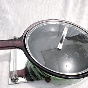 May include: A vintage green glass casserole dish with a clear glass lid. The lid has a silver handle and etched floral designs. The dish has a dark purple handle and rim. The casserole dish is on a mirrored surface.