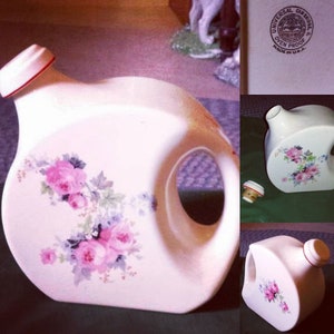 May include: White ceramic pitcher with a floral design featuring pink and blue flowers. The pitcher has a unique shape with a cut-out handle and a red lid. The pitcher is labeled "Universal Can Bridge Oven Proof Made in U.S.A."