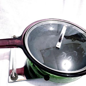 May include: A round purple glass skillet with a matching clear glass lid. The lid features a delicate etched floral pattern and a central silver-toned metal handle. The skillet has a long, integrated purple glass handle extending to the left.