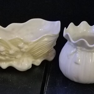 May include: Two white ceramic planters, one shaped like a lily of the valley flower and the other shaped like a bag with a bow. Both planters have a ruffled rim.