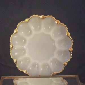 May include: A white, scalloped egg serving dish with a gold-colored trim. The dish has 12 individual egg compartments and a central area. The dish is likely made of glass or ceramic and is designed for serving eggs.