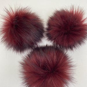 May include: Three burgundy and black faux fur pom-poms. Each pom-pom is round and fluffy, with long, spiky strands extending outwards. These decorative items are suitable for various craft projects, adding a touch of texture and volume to accessories.