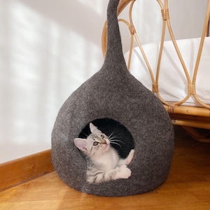 K&ouml;nnte beinhalten: Ein graues Katzenbett aus Filz in Tropfenform mit einer kleinen &Ouml;ffnung. Ein kleines grau-wei&szlig;es Tabby-K&auml;tzchen schaut aus der &Ouml;ffnung. Das Katzenbett hat eine lange, spitze Oberseite und steht auf einem Holzboden.