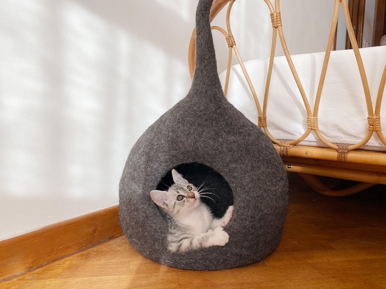 K&ouml;nnte beinhalten: Ein graues Katzenbett aus Filz in Tropfenform mit einer kleinen &Ouml;ffnung. Ein kleines grau-wei&szlig;es Tabby-K&auml;tzchen schaut aus der &Ouml;ffnung. Das Katzenbett hat eine lange, spitze Oberseite und steht auf einem Holzboden.