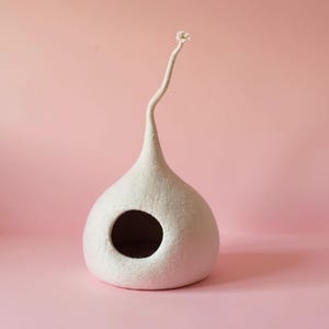 Può includere: Una cuccia per animali in lana infeltrita bianca con una base rotonda e bulbosa e un'apertura circolare sul davanti. La struttura si restringe verso l'alto in un collo lungo, sottile e curvo che termina con una piccola e delicata forma a fiore.