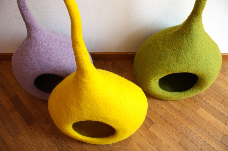 May include: Three felt cat beds in different colors: purple, yellow, and green. The beds are shaped like a teardrop with a large opening at the bottom.