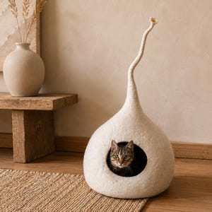 Può includere: Una cuccia per gatti in lana infeltrita color crema a forma di goccia con un lungo stelo sottile e ricurvo sulla parte superiore. La struttura presenta un'apertura circolare sul davanti, che rivela un gatto tigrato all'interno.