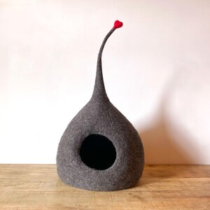 Pode incluir: Uma cama para gatos de feltro cinza com uma cauda longa, pontiaguda e com ponta vermelha. A cama tem uma abertura redonda para o gato entrar.