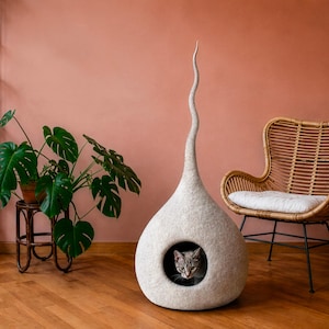 K&ouml;nnte beinhalten: Ein modernes Katzenbett in hellem Grau mit einer hohen, geschwungenen Form. Das Bett hat eine runde &Ouml;ffnung, in der eine graue Tabby-Katze zu sehen ist. Ein Topf mit einer Pflanze und ein Korbstuhl sind im Hintergrund.